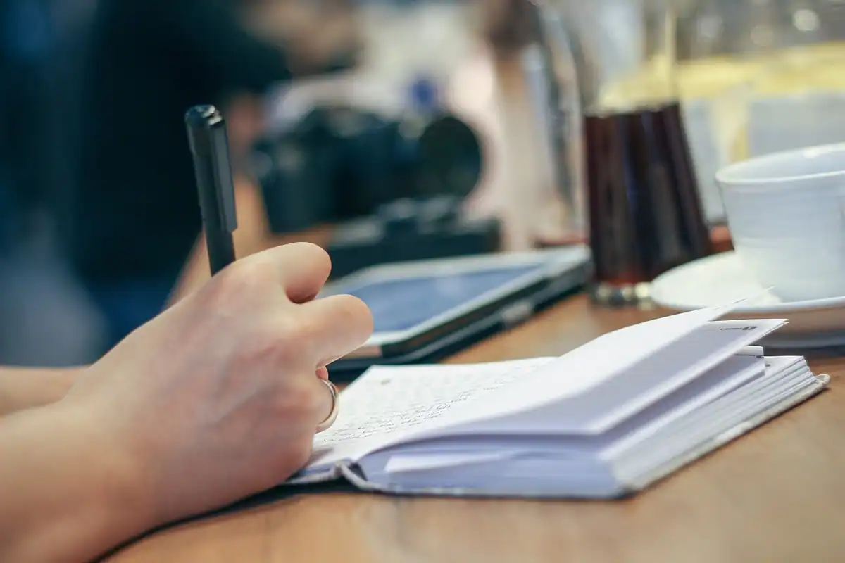 Mână care notează cu pixul într-un carnet, la masa din cafenea