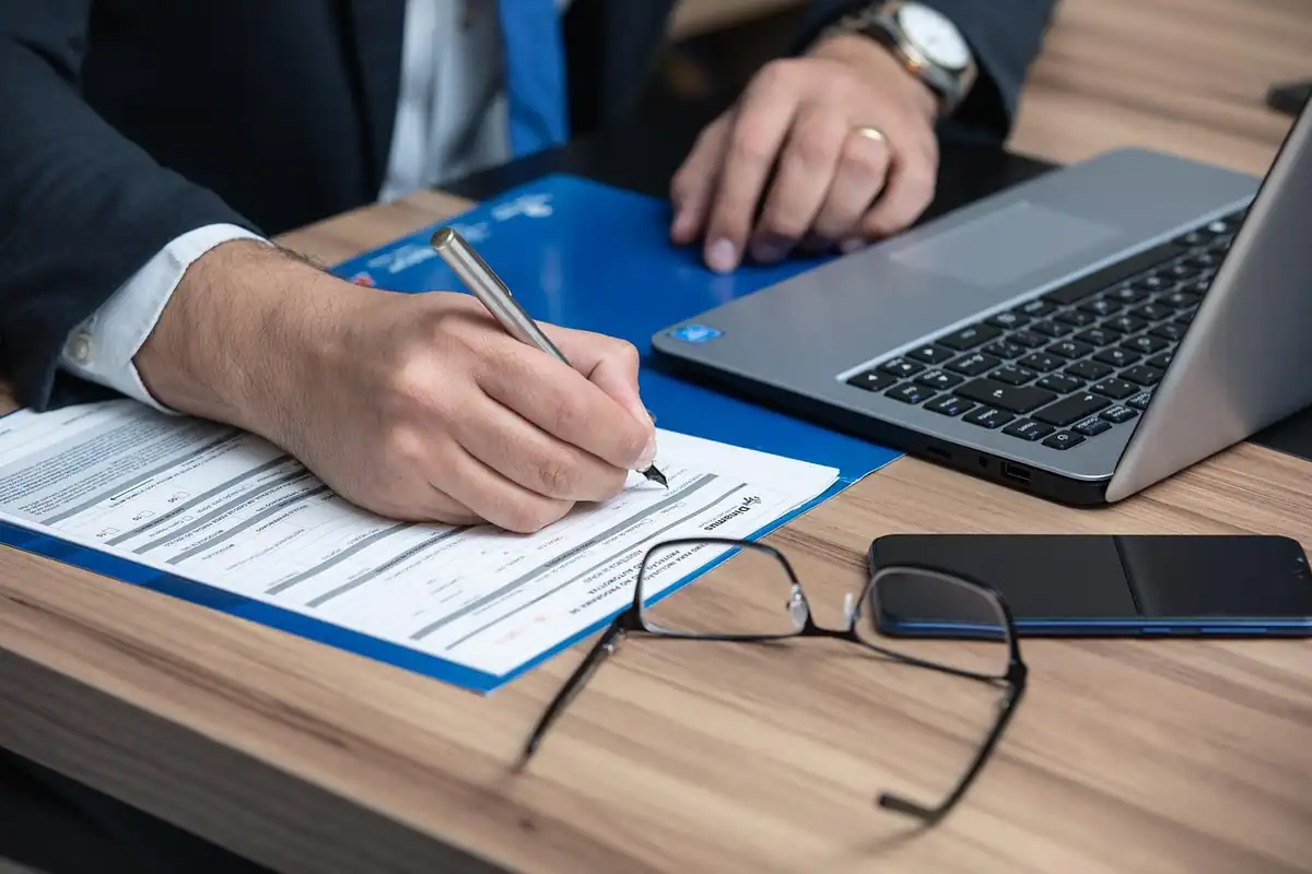 Mână care completează un formular lângă laptop și ochelari, la încheierea contractului