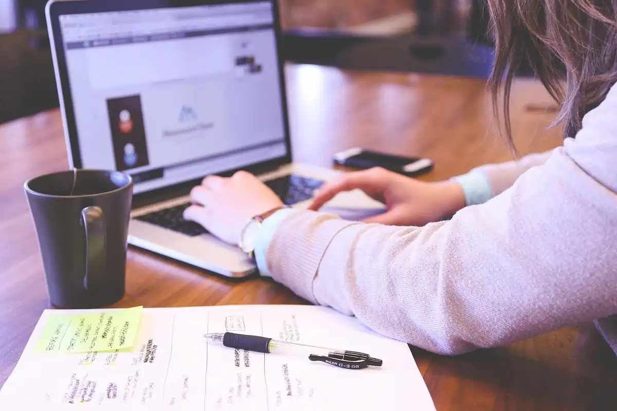 Femeie care lucrează la laptop, cu hârtii pe birou, la completarea registrului