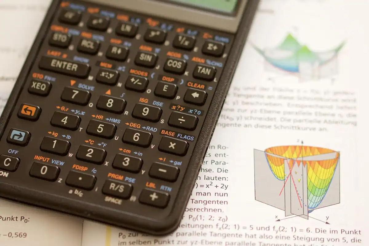 Mâini care scriu pe hârtie la un birou, cu calculator, caiet și planuri desenate