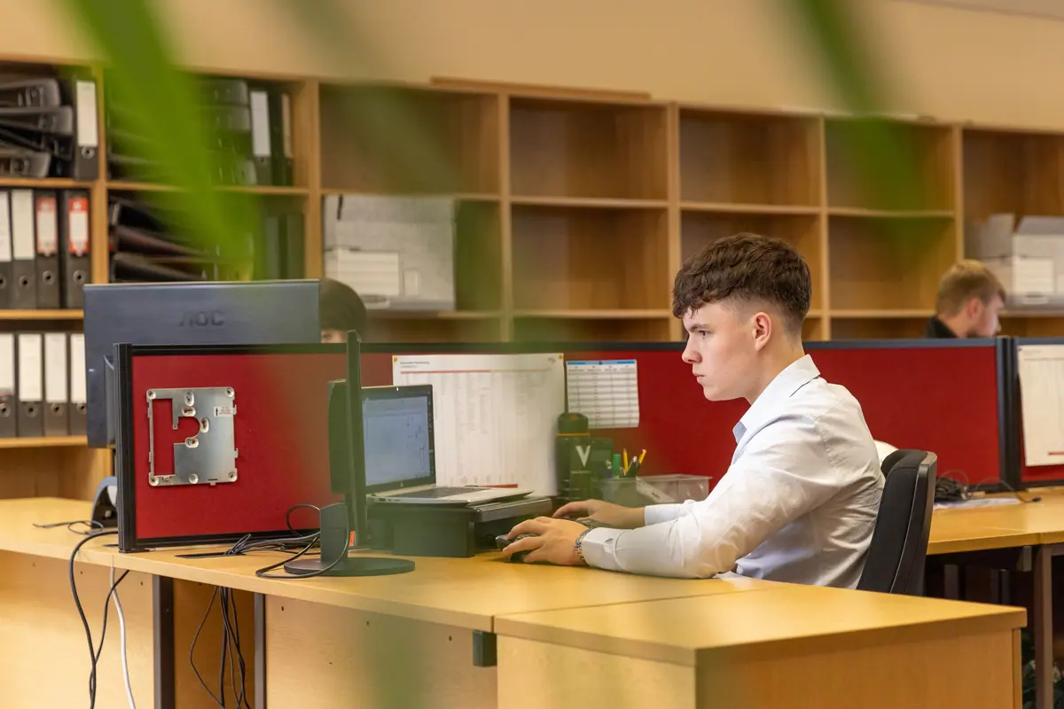 Tânăr care lucrează la computer într-un birou deschis, cu rafturi de dosare